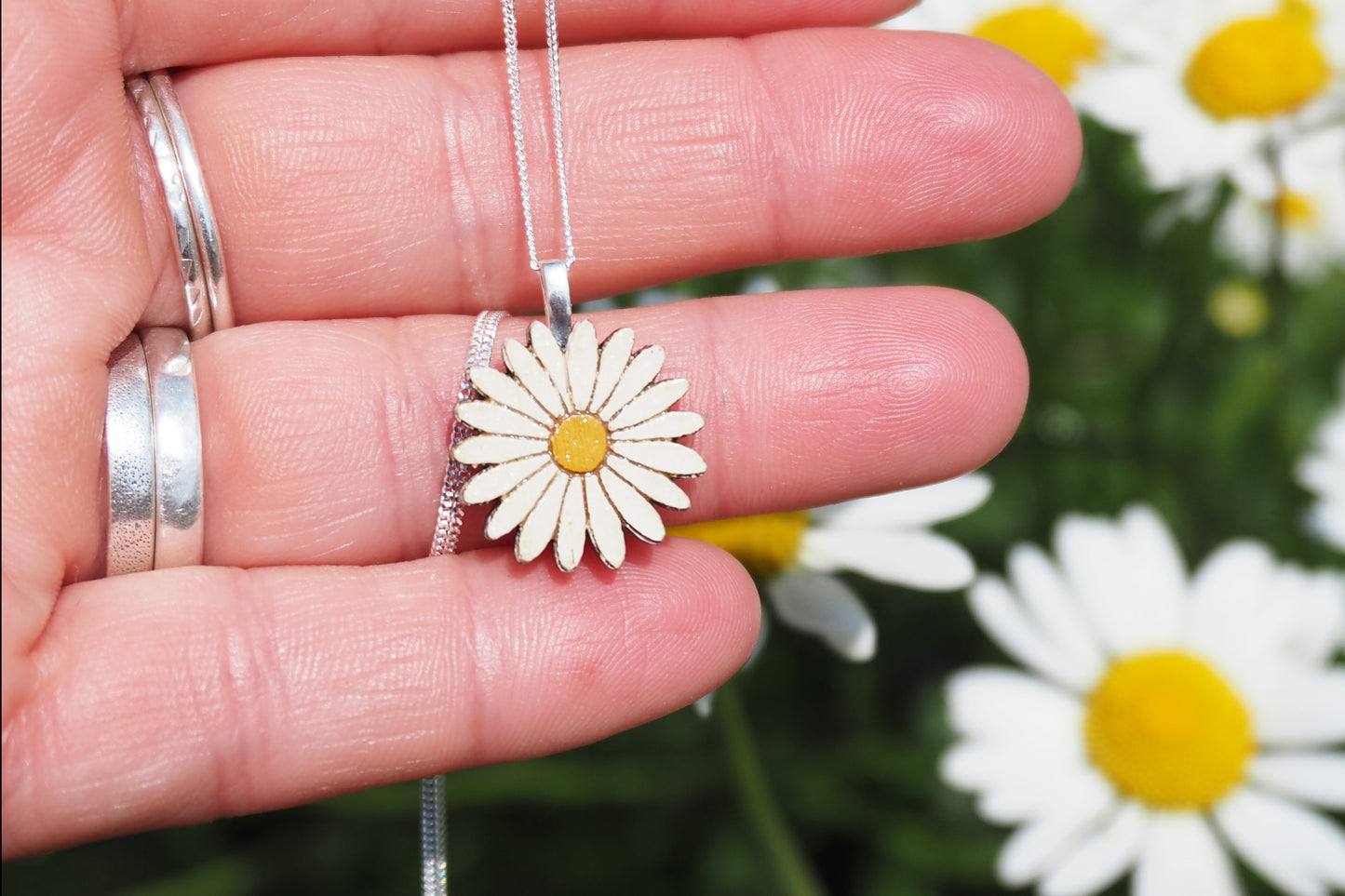 Silver daisy flower necklace held in a hand with flowers in the background