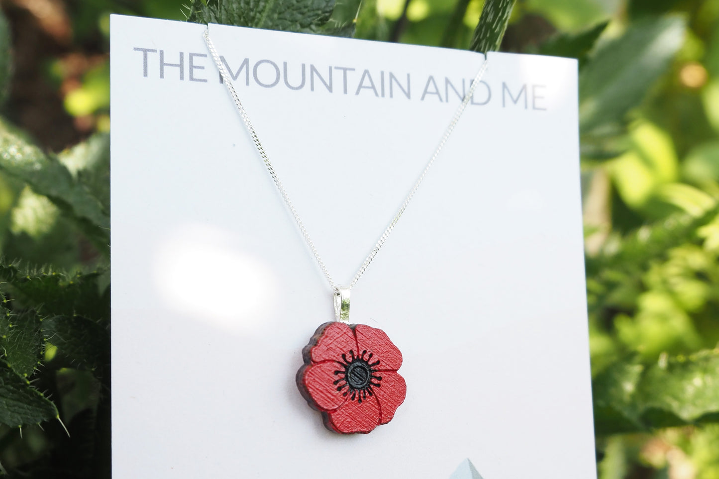 Necklace with a red poppy flower pendant on a card with 'The Mountain and Me' text, against a green leafy background.